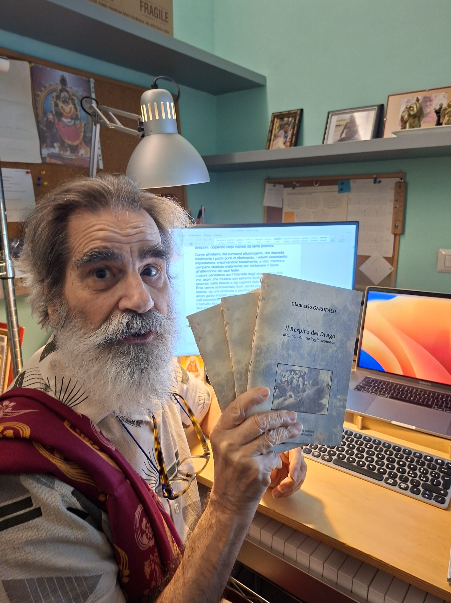 Persona con barba e capelli grigi, seduta davanti a computer con libri e foto sullo sfondo, tiene in mano un libro di YogaLila